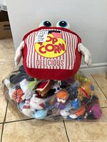 Full lot image showing large plush popcorn bag on top of a plastic bag filled with various plush toys including characters in multiple shapes and colors.