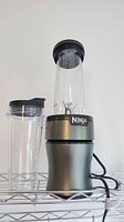 Ninja blender base with tall blending cup attached, showing blade assembly and single-serve blending cup with lid next to it on wire shelf.