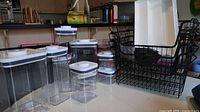 Full set of 7 varying sized transparent plastic canisters with white lids, 2 black wire baskets, and 1 white cutlery tray arranged on white surface near kitchen counter.