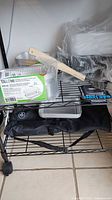 Pampered Chef BBQ tool set stored on black shelving unit with black carrying case, disposable aluminum food packaging containers, magnetic gas level indicator, and wooden-handled tools.