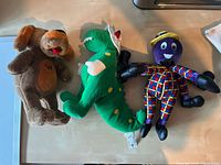 Photo showing three plush toys: a brown dog, a green alligator, and a purple octopus, all placed side by side on a table.