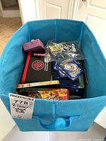 Blue fabric bin containing loose English Pokemon cards, a pink card holder, Pokemon and Yu-Gi-Oh! metal tins, and loose accessories in plastic bag.