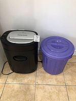 Black and silver Royal 1212X paper shredder next to a purple plastic trash can on tiled floor.