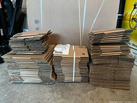Front view of four stacks of flattened small Staples shipping boxes, bundled with white straps, placed on a concrete floor against a cardboard background.