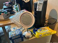 Photo showing tower fan, dishwasher cleaning boxes, lighted makeup mirror, electromagnetic aromatherapy diffuser, and Lysol wipes on tabletop.