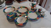 Full set displayed on table showing plates, bowls, cups, and teapot with colorful geometric pattern around the rim/body.