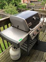 Front-left angle of grill on deck showing propane tank, side shelf, wheels and overall exterior.