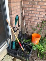 Photo showing Yardworks electric grass trimmer, multiple hand tools including rakes and shovels, and orange bucket placed in black crates near a brick wall.