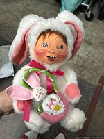 White and pink plush toy dressed as a bunny, holding a pink heart decoration with ribbons, showing detailed face and costume.
