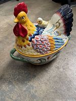 Side view of the lidded porcelain tureen shaped like a colorful rooster with a chick figurine on the lid, showcasing the detailed hand-painted design and green handles.