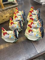 Photo showing eight colorful ceramic napkin rings shaped like roosters with chicks, arranged in two rows on table.