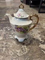 Full view of white porcelain teapot with pink and purple floral patterns and gold handle on a concrete floor.
