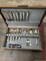 Silver-plated flatware partial set inside original case showing multiple forks, spoons, knives, and serving utensils organized in padded compartments.