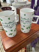 Two tall white ceramic mugs with green shamrocks and gold trim, square handles, placed on wooden surface.