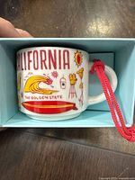 Starbucks ceramic mug ornament with California graphics and red hanging string in light blue box.