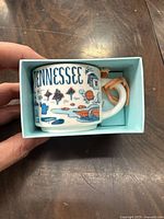 Top view of the Tennessee mini mug ornament inside light blue box, showing colorful Tennessee-themed illustrations and hanging string loop on handle.