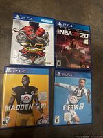 Four PlayStation 4 game cases and discs: Street Fighter V, NBA 2K20, Madden NFL 19, FIFA 19 on a brown carpet background.