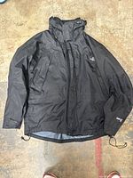 Full black men's jacket laid out showing hood, length, and button/zipper closure.