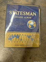 Cover photo of Statesman Deluxe Stamp Album in blue and gold with world globe image and map motifs, showing it is a large deluxe album designed for extensive stamp collections.