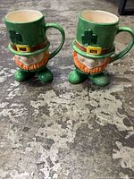 Two ceramic mugs shaped like leprechauns, front angle showing detailed faces, green hats with shamrock, orange beards, and small feet bases.
