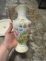 Close-up of vintage white porcelain vase with multi-colored floral pattern and gold-tone handles, showing detailed design and finish.