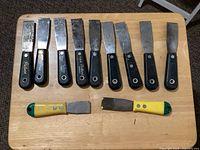 Photo showing 11 putty knives and tools laid out in a row on a wooden surface, including 9 with black handles and 2 with yellow/green handles, blades show signs of use.