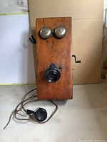 Front view of antique wooden wall telephone with metal bells, mouthpiece and wired black earpiece.