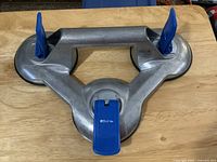 Top view of the triangular aluminum suction lifter showing three blue suction cup activation levers, placed on a wood surface.