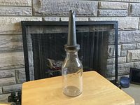 Front view of clear glass oil bottle standing on a wooden table, showing metal pour spout and general shape.