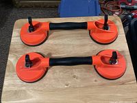 Two new glass lifting suction cups placed on a wooden surface showing top view: orange handles with black rubber grips and levers for suction locking.