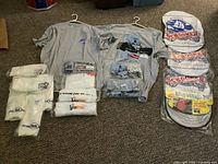 Wide view of all items including multiple folded T-shirts, two grey T-shirts on hangers, car steering wheel cover, and inflatable beer cans.