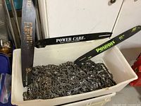 Bin filled with approximately 15-20 metallic chainsaw chains piled together showing some use wear. Three chainsaw guide blades rest on the edge, one marked Power Care.