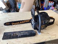Left side view of Poulan Pro chainsaw on wooden surface, showing 16 inch bar with chain and protective cover next to it