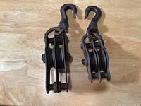 Top view of two vintage cast iron double pulleys with hooks placed side by side on a wooden surface, showing the pulleys and hooks clearly.