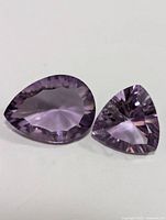 Two faceted amethyst gemstones, one pear-shaped and one trillion-shaped, placed side by side on a white surface.