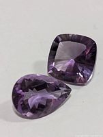 Close-up of two faceted amethyst gemstones on a white surface, one pear-shaped and one cushion-cut.