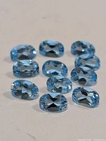 Close-up photo of 11 oval faceted blue topaz gemstones arranged on a white surface, showing clear light blue color and crystal clarity.