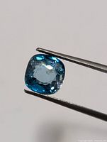 Close-up photo of the cushion-cut bright blue zircon held in tweezers against a plain background showing the cut and clarity.