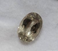 Close-up photo of the oval faceted color changing Zultanite gemstone lying on a white cushion surface, showing the light yellowish-green hue.