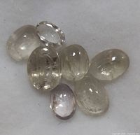 Seven oval cabochon cut transparent gemstones showing internal inclusions placed on plain white background.