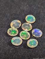 Eight oval-cut faceted genuine opal gemstones displayed on black fabric. Showcasing their translucent quality and bright play-of-color with green, blue, and orange flashes.