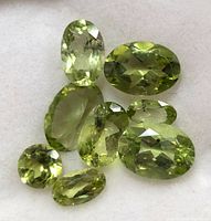 Photo showing eight oval and round faceted light green peridot gemstones arranged closely on a light background.