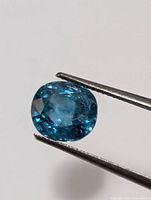 Close-up image of a blue zircon gemstone held by tweezers showing its cushion cut and faceted surface.