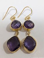 Front view of two purple faceted amethyst stones in gold-tone bezels on hook earrings.