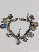 Photo of silver charm bracelet with visible detailed charms including butterfly, blue enamel church medallion, heart with inscription, crosses, lion figure, and other charms on chain.