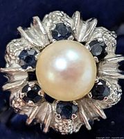Close-up image of the front of the ring showing the central pearl surrounded by six blue sapphire stones, with detailed silver floral motifs.