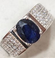 Close-up image of a sterling silver ring with an oval deep blue synthetic sapphire stone and multiple surrounding small clear cubic zirconia stones on the band.