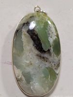 Close-up image of an oval pendant with a polished gemstone that has green, white, and black patterning set in a silver frame.