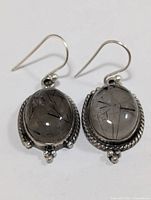 Pair of silver earrings with oval-shaped rutilated quartz stones showing black needle-like rutile inclusions inside. Silver frames have twisted rope border and small balls decoration with hook wires for ears.