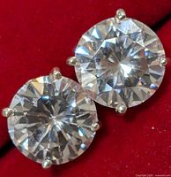 Close-up photo of two silver stud earrings with clear round cubic zirconia stones in four-prong settings on red background.